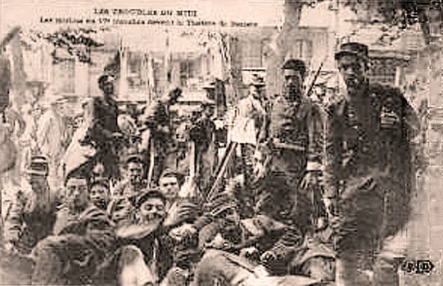 Carte postale sépia : des soldats en uniforme et képi, fusils en main, entourés de civils, posent assis et debout devant un bâtiment.