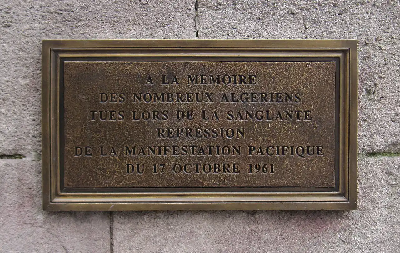 Plaque de bronze rectangulaire fixée sur un mur de pierre, portant une inscription en lettres capitales.