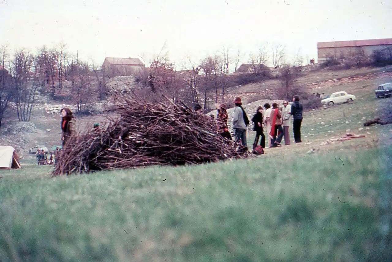 Photographie en couleur d'un groupe de personnes debout dans un pré à côté d'un grand tas de branchages, avec des fermes et des voitures en arrière-plan.