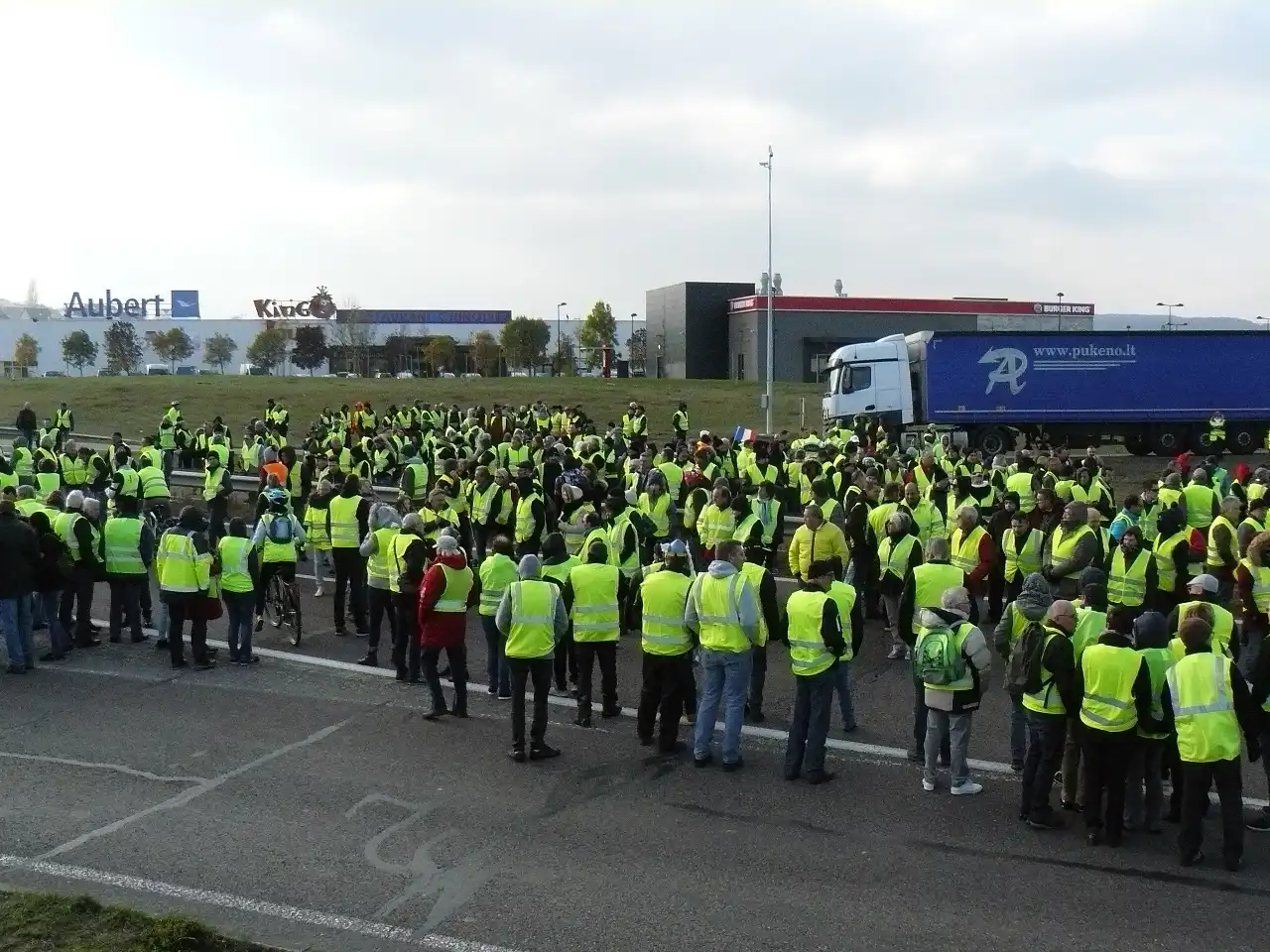 Plusieurs centaines de personnes en gilet jaune rassemblées sur une route à quatre voies, devant une zone commerciale ; un camion est arrêté à droite.