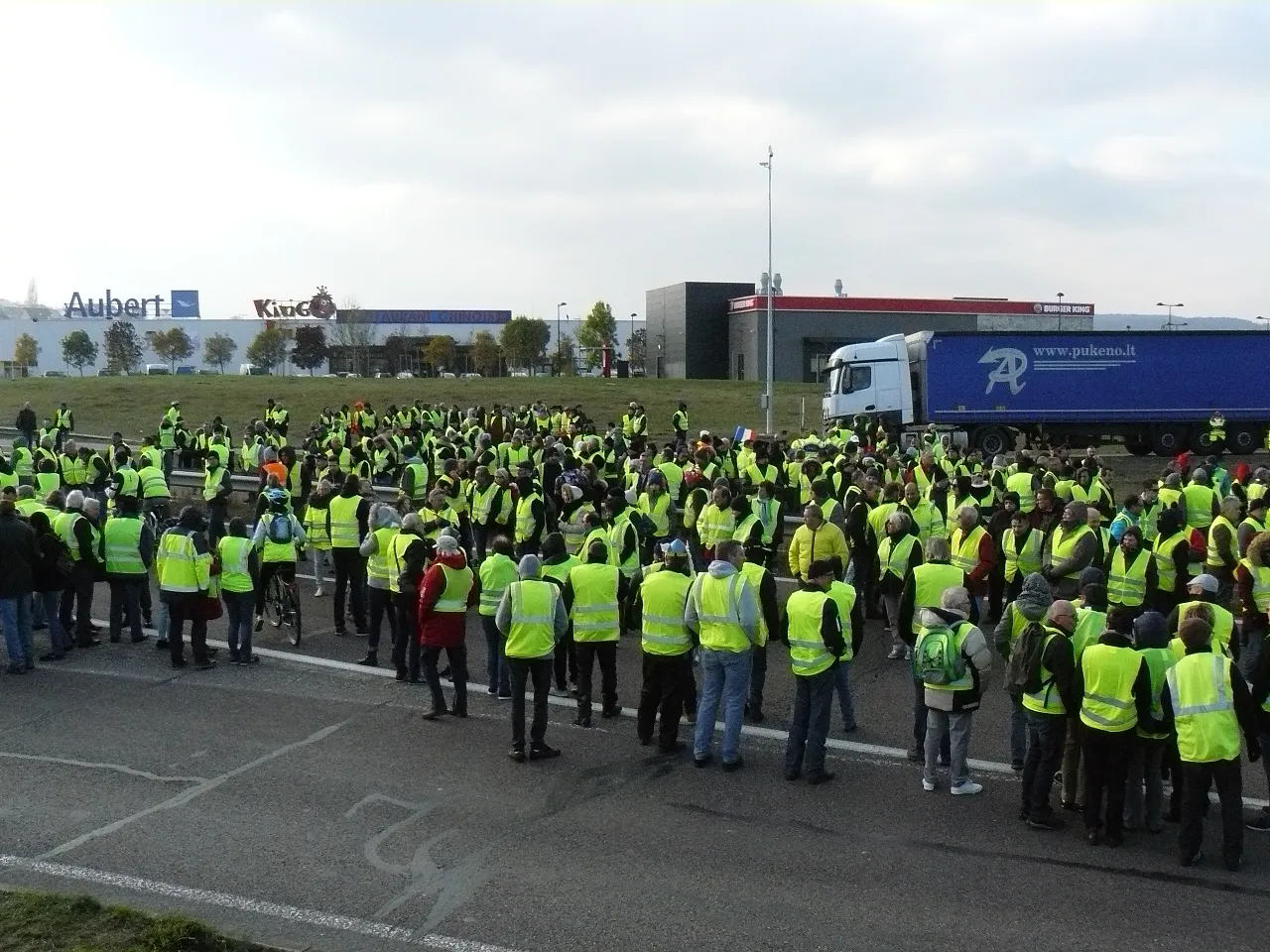 Plusieurs centaines de personnes en gilet jaune rassemblées sur une route à quatre voies, devant une zone commerciale ; un camion est arrêté à droite.